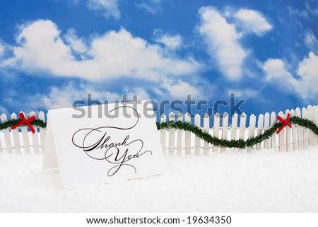 Thank you card sitting on snow with white fence and green garland on a sky background