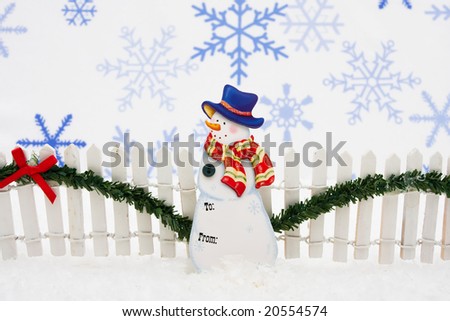Snowman gift tag with white fence and green garland on snowflake background