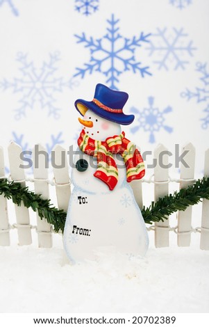 Snowman gift tag with white fence and green garland on snowflake background, Snowman