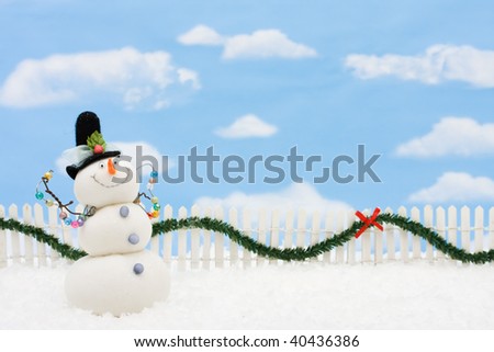 A snowman on a white picket fence with garland on a  sky background