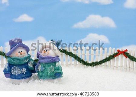 Two snowmen on a white picket fence with garland on a  sky background, gingerbread house