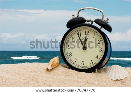 An alarm clock with seashells sitting on the sand with water, vacation time