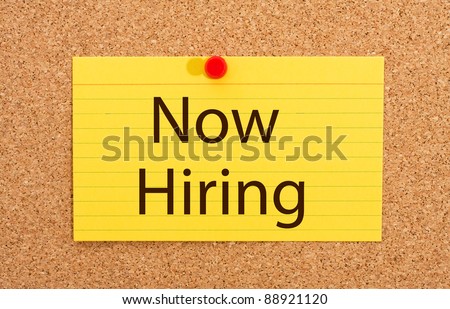 A yellow index card on a cork board with the words now hiring on it