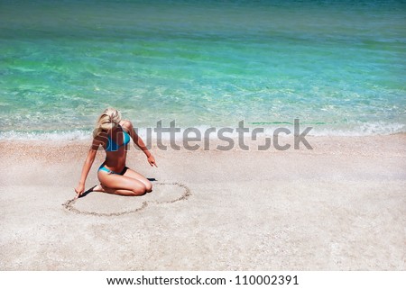 beautiful slender young woman painting the heart on the sea sand in summer bright day