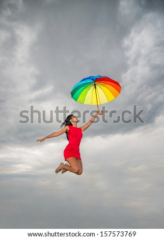 Young pretty woman in red dress jumping with rainbow umbrella against overcast sky