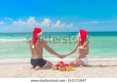 Lovers couple in red santa hats and many golden packed presents on sea beach - christmas vacation concept