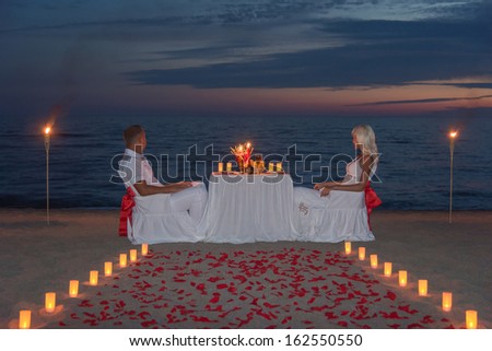 Young couple share a romantic dinner with candles, torches and way or rose petals on sea sandy beach against sunset - wedding day, proposal of marriage or honeymoon concept