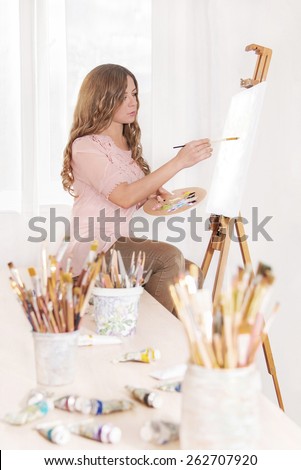 Cute woman artist painting in oils on canvas with brushes in her workshop near window