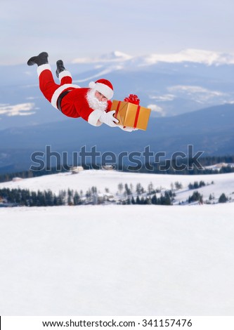 Santa Claus flying fall over with big golden gift box above snowy winter landscape (houses, mountains, fir trees forest), Happy New Year and Merry Christmas coming concept