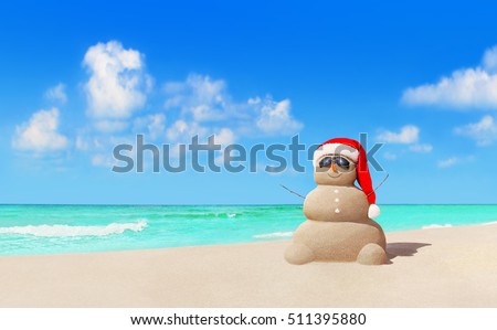 Positive sandy Snowman in red Christmas Santa Claus hat and sunglasses at sunny tropical beach. New Year concept for travel destinations in hot tourist countries