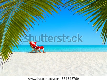 Christmas Santa Claus relaxing on sunlounger at ocean sandy tropical beach under palm leaves. Happy New Year travel destinations to hot countries concept