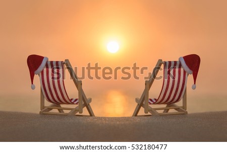 Two striped red-white deckchairs with red Santa hats for romantic couple at ocean sandy beach during sunset. Happy New Year and Merry Christmas travel destinations to hot countries concept