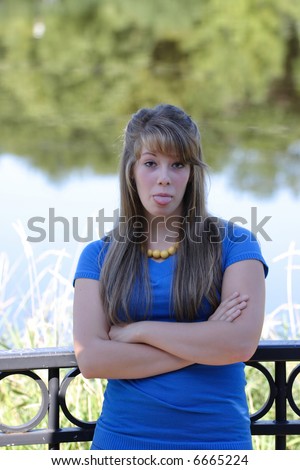A pretty teenage girl sticking her tongue out while standing against a black cast iron railing near a river front.