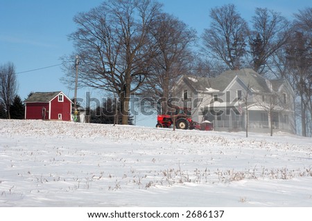 Chore after a storm, blowing out the driveway on a farm with a tractor mounted snowblower. Snow from blower forms a cloud in front of the house