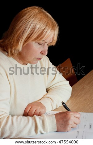 Senior woman sitting at the table and filling in an official document (on dark background)