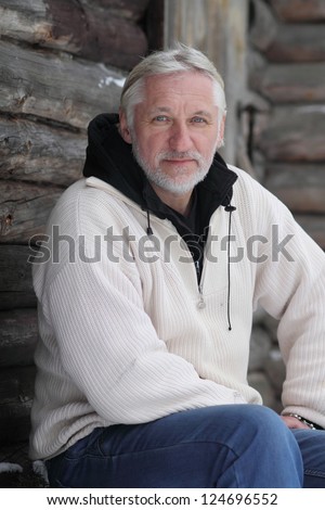 The attractive gray-haired elderly  man sits beside log home