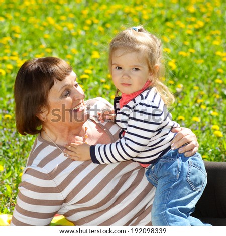Grandmother and granddaughter having fun on a blanket on a background of yellow flower meadows. Naughty beautiful little baby with her grandmother outdoors. Grandmother and grandchild