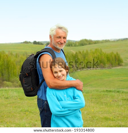 Portrait of grandfather with a travel backpack and his granddaughter on background a spacious landscape. Grandfather and granddaughter travel to natural places