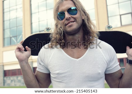 muscular long-haired guy in sunglasses with skateboard. Urban scene. Outdoor lifestyle portrait