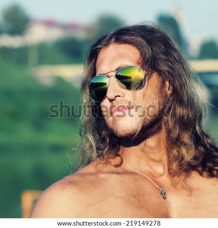 Portrait courageous unshaven man with long red hair and a cross on his neck on background of sky.