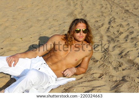 Fitness model man with long hair and wearing sunglasses posing on the beach