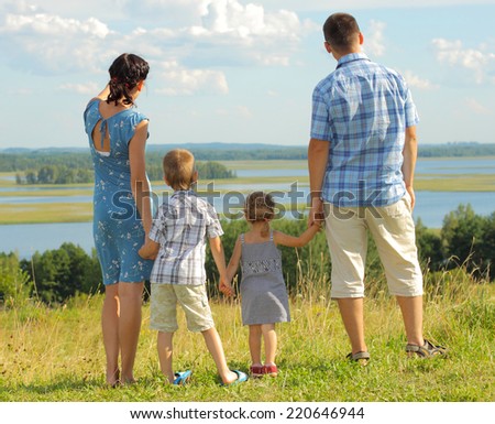 happy young family of four having fun on the top of a hill on a background of clouds. family of four standing on a hill and looking at spaces. View from the back.