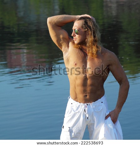 Beautiful long-haired man with a naked muscular torso walks on the beach. Fitness model man in sunglasses on the beach near the water.