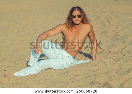 Fitness model man with long hair and wearing sunglasses posing on the beach