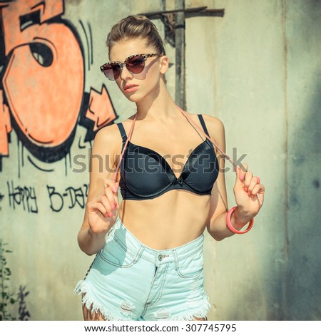 Stylish fashionable hipster blonde in sunglasses with a pink belt posing on wall background