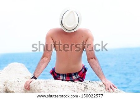 Man on beach lying on beach rocks wearing hipster summer hat. Young male model enjoying summer travel holiday by looking at the ocean