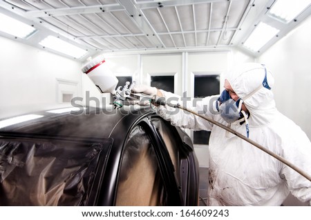 Auto Painter Spraying A Car Body With Black Paint And Protective Gear