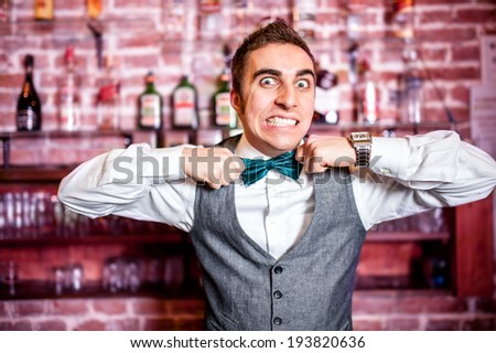 Portrait of angry and stressed bartender or barman with bowtie behind the bar with alcoholic drinks around
