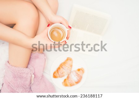 close-up of woman legs in bed, with fluffy slippers drinking a coffee and reading a good book. Woman relaxation with coffee, croissants and a good reading book