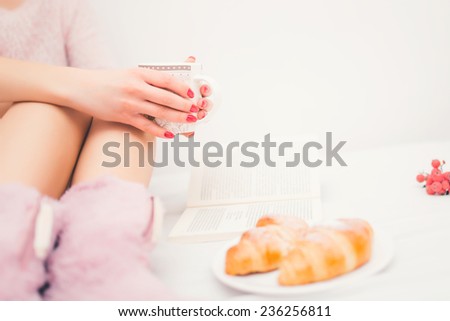 close-up of woman legs in bed, with fluffy slippers drinking a coffee and reading a book. Woman relaxation with coffee, croissants and breakfast in bed