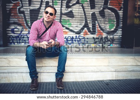 Portrait of handsome man with phone in hand, casually dressed with shirt, jeans and sunglasses against graffiti painted wall. Stylish european man traveling and taking pictures with mobile phone