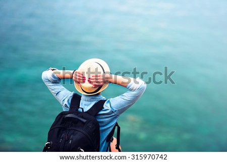 young woman on summer vacation, hiking on coastline and staring at sea wearing hat and backpack. Travel and adventure concept
