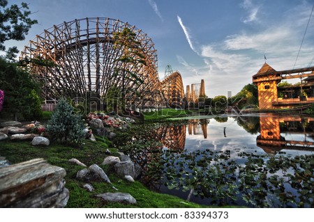 Side view of a wooden roller coaster being mirrored in a lake with blue, clear sky