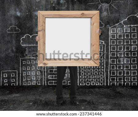 businessman holding wooden frame blank whiteboard with sun clouds buildings doodles wall on dark mottled concrete floor background