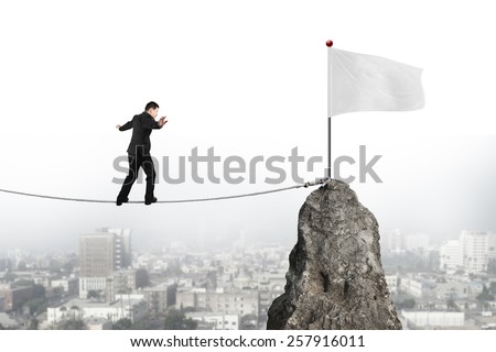 Businessman walking and balancing on rope toward white flag of mountain peak with cityscape background