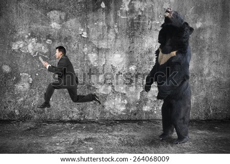 Businessman holding tablet running from an angry bear with gray mottled concrete wall and floor background