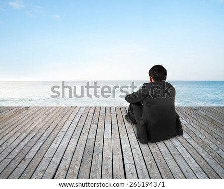 Rear view of black suit businessman with hands holding his knee sitting on wooden floor and sky sea background