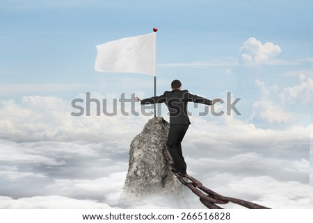 Businessman walking and balancing on old iron chain toward white flag of mountain peak with cloudscape background