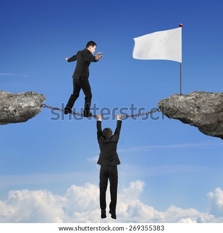 Businessman balancing on the broken rusty iron chains another man holding connect two cliffs and walking toward white flag with sky cloudscape background, business teamwork concept.
