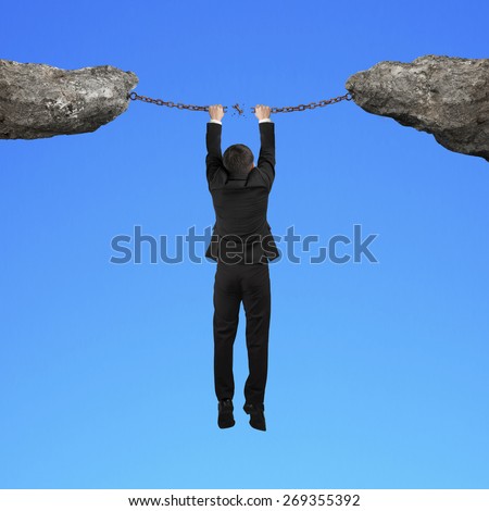 Businessman hand hanging on cracking rusty iron chains connect two cliffs with blue background