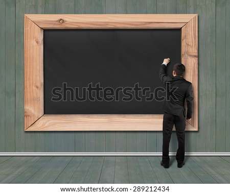 Businessman drawing on blank black chalkboard with dark green wood wall and floor background