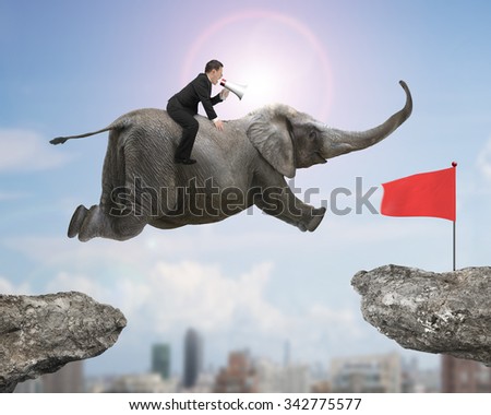Man with using speaker riding elephant flying toward red flag on cliff, with sunny sky cityscape background.