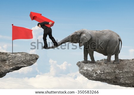 Businessman carrying red arrow up sign balancing on the nose of elephant toward red flag on cliff, with sky background.