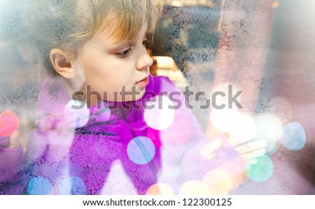Little blond girl beyond frozen shop window glass with colorful blurred lights