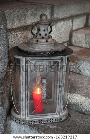 Old metal outdoor lamp with red burning candle stands on stone stairs