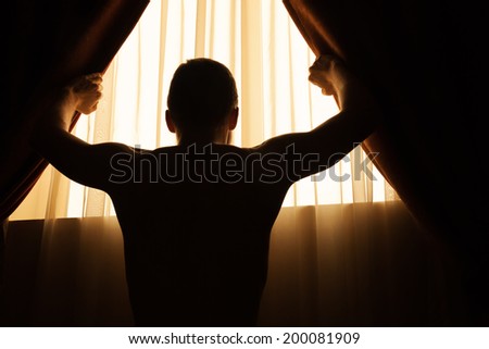 Man in dark room opens curtains on window to the morning light